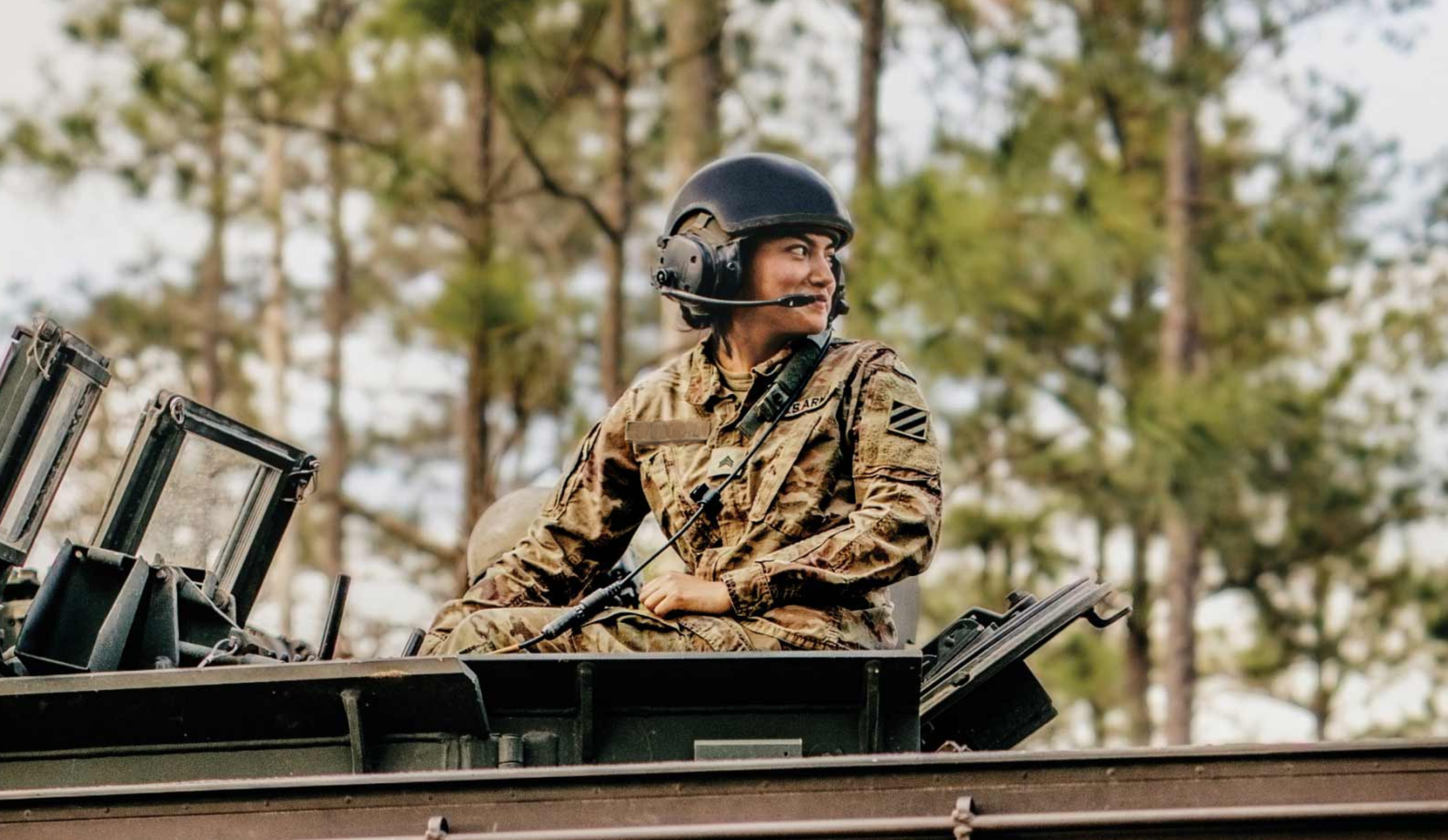 A female soldier in a camouflage uniform and tactical headset helmet sits atop a military vehicle, looking to the side. She has a focused expression, and the vehicle appears to be an armored personnel carrier or tank. The background consists of a blurred forest with tall trees, suggesting a training or deployment environment.